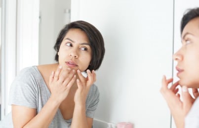 a woman looking in the mirror popping a pimple on her chin