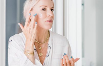 Renee Rouleau standing infront of a mirror and applying products on her face for healthy aging